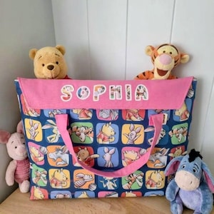 Può includere: Una borsa tote rettangolare in tessuto con un motivo a griglia ripetuto di personaggi di Winnie the Pooh su sfondo blu scuro. La borsa presenta una patta superiore rosa con il nome SOPHIA in lettere colorate e due manici in tessuto rosa.