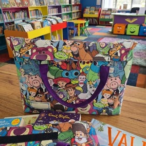 Pu&ograve; includere: Una borsa in tessuto con una stampa multicolore di vari personaggi animati del franchise Toy Story. La borsa presenta manici in tessuto viola e un'etichetta bianca rettangolare sul davanti. &Egrave; posizionata su una superficie in legno.