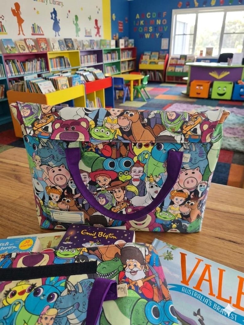 Pu&ograve; includere: Una borsa in tessuto con una stampa multicolore di vari personaggi animati del franchise Toy Story. La borsa presenta manici in tessuto viola e un'etichetta bianca rettangolare sul davanti. &Egrave; posizionata su una superficie in legno.