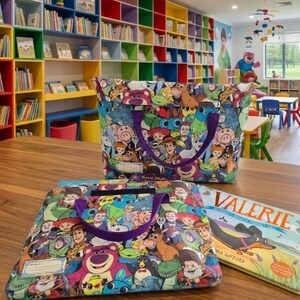Pu&ograve; includere: Due borse in tessuto con una stampa colorata di vari personaggi di film d'animazione, tra cui un orso rosa, un cowboy e un ranger spaziale. Ogni borsa ha manici viola e una piccola etichetta rettangolare bianca per scrivere un nome.