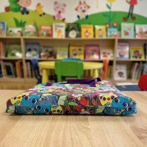 Pu&ograve; includere: Una borsa in tessuto piatta e rettangolare caratterizzata da una vivace stampa multicolore con vari personaggi dei cartoni animati in tonalit&agrave; di blu, rosso, verde e giallo. La borsa ha due manici in tessuto viola sul bordo superiore.