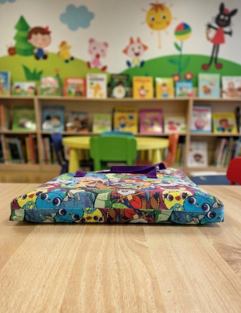 Pu&ograve; includere: Una borsa in tessuto piatta e rettangolare caratterizzata da una vivace stampa multicolore con vari personaggi dei cartoni animati in tonalit&agrave; di blu, rosso, verde e giallo. La borsa ha due manici in tessuto viola sul bordo superiore.