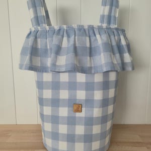 May include: A cylindrical fabric storage basket featuring a light blue and white gingham check pattern. The basket has two upright fabric handles and a decorative ruffled trim around the top edge. A small square wooden label with a kangaroo logo is centered.