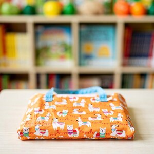 May include: A rectangular fabric pouch featuring an orange background with a repeating pattern of white llamas wearing colorful patterned blankets and saddles, accented by red string lights. The pouch has light blue fabric handles attached.