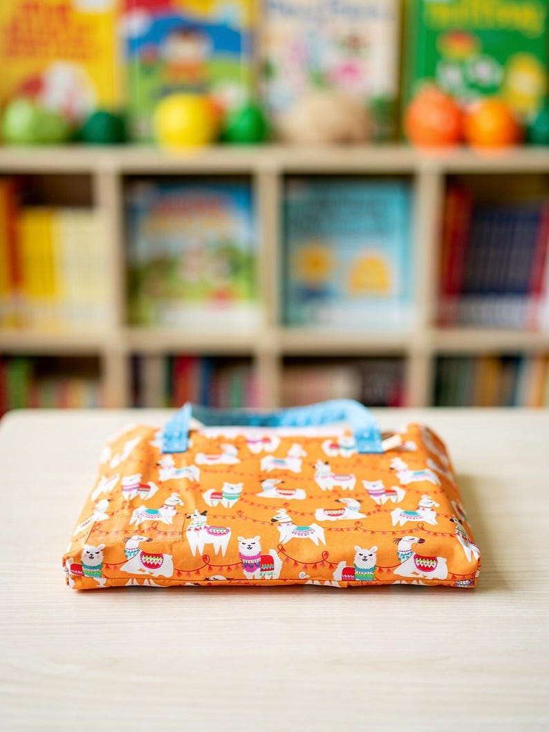 May include: A rectangular fabric pouch featuring an orange background with a repeating pattern of white llamas wearing colorful patterned blankets and saddles, accented by red string lights. The pouch has light blue fabric handles attached.