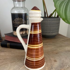 May include: A brown and white ceramic oil cruet with a handle and a spout. The cruet features horizontal stripes in varying shades of brown and yellow. The top is white with a brown and yellow conical stopper. The cruet is on a wooden surface.