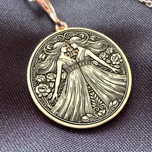 Peut inclure: Un collier pendentif doré avec un motif gravé de deux femmes en robes fluides, entourées d'éléments floraux. Le pendentif est suspendu à une chaîne délicate, sur un fond de tissu sombre.