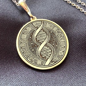 Puede incluir: Un collar con colgante dorado con un diseño detallado de doble hélice de ADN dentro de un marco circular. El colgante cuelga de una delicada cadena plateada. El collar se muestra sobre una tela azul oscuro.