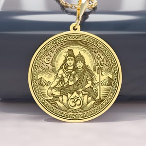 May include: A gold-colored pendant necklace featuring a detailed engraved image of two figures in a seated pose, surrounded by ornate patterns. The pendant hangs from a delicate gold chain. The pendant is round with a detailed design.