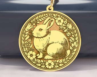 May include: A gold-colored pendant necklace featuring a detailed illustration of a rabbit within a floral wreath. The pendant hangs from a delicate gold chain. The rabbit is depicted in a seated position, with intricate details. The background is a dark blue surface.