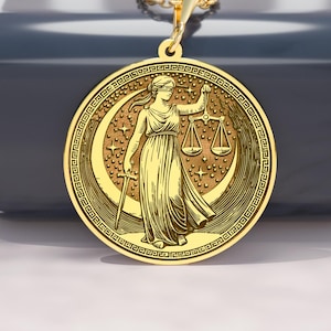 Puede incluir: Un collar con colgante dorado que presenta una detallada representación de la Dama de la Justicia, sosteniendo una balanza y una espada. El colgante redondo está suspendido de una delicada cadena dorada. El diseño incluye una luna creciente y estrellas, con un borde de grecas.