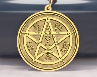 May include: Gold-colored pendant necklace featuring a pentagram design with intricate symbols and text. The pendant hangs from a delicate gold chain. The necklace is displayed on a dark blue surface.
