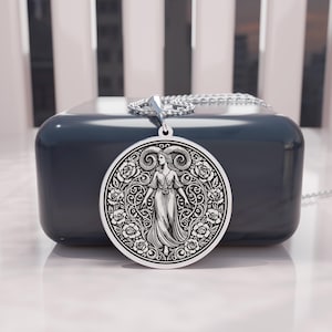 May include: A silver pendant necklace featuring a detailed engraving of a woman with ram horns, surrounded by floral patterns. The pendant hangs from a delicate chain. The design is set against a dark blue background.