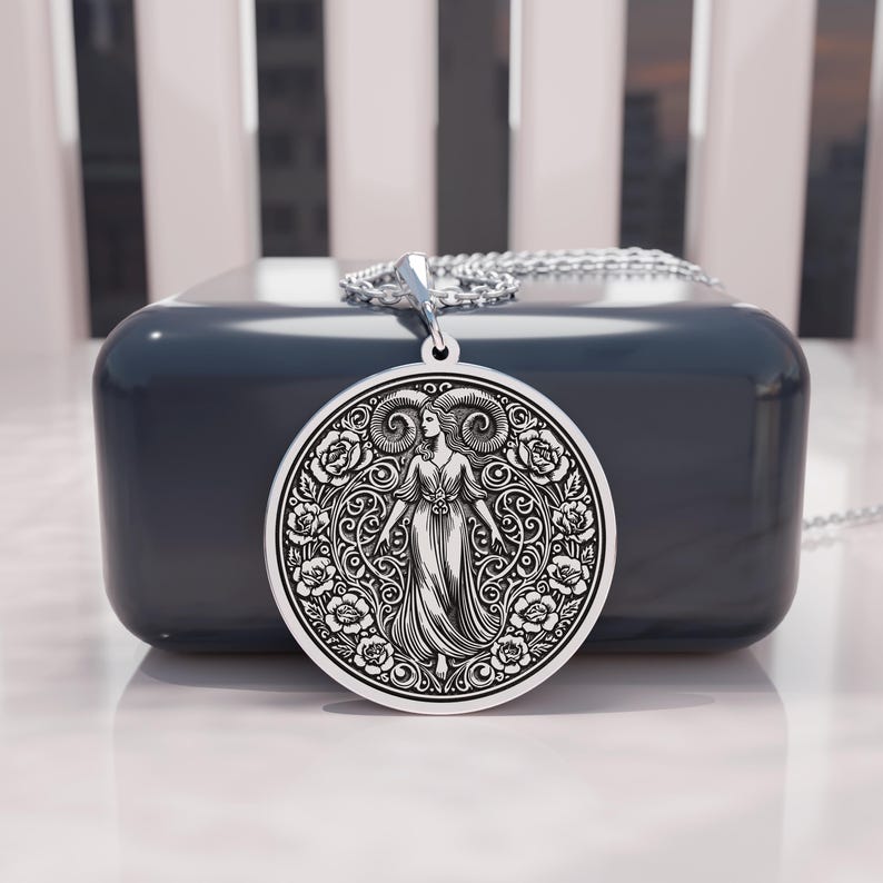 May include: A silver pendant necklace featuring a detailed engraving of a woman with ram horns, surrounded by floral patterns. The pendant hangs from a delicate chain. The design is set against a dark blue background.