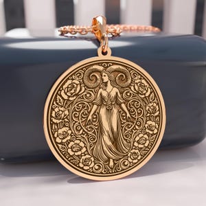 May include: A gold-colored pendant necklace featuring a detailed circular design. The pendant showcases a woman figure surrounded by roses and ornate scrollwork. The necklace has a delicate chain and a decorative bail.