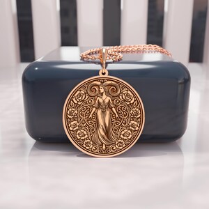 May include: A gold-colored pendant necklace featuring a detailed Aries zodiac design. The round pendant showcases a woman with ram horns, surrounded by floral patterns. The necklace is displayed on a dark blue surface, with a gold chain.