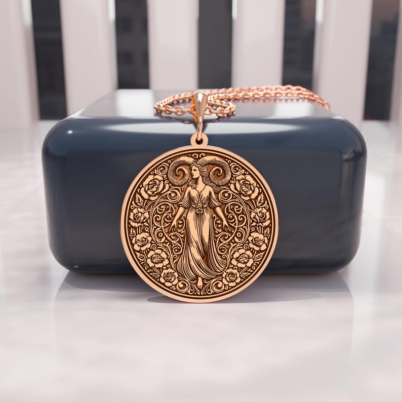 May include: A gold-colored pendant necklace featuring a detailed Aries zodiac design. The round pendant showcases a woman with ram horns, surrounded by floral patterns. The necklace is displayed on a dark blue surface, with a gold chain.