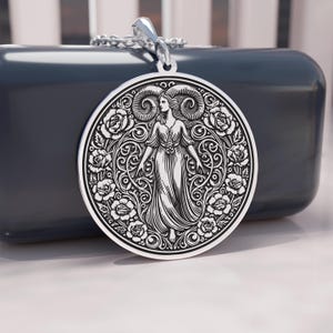 May include: A silver pendant featuring a detailed engraving of a woman with ram horns, surrounded by roses and ornate scrollwork. The pendant hangs from a silver chain. The design is circular and has a vintage aesthetic.