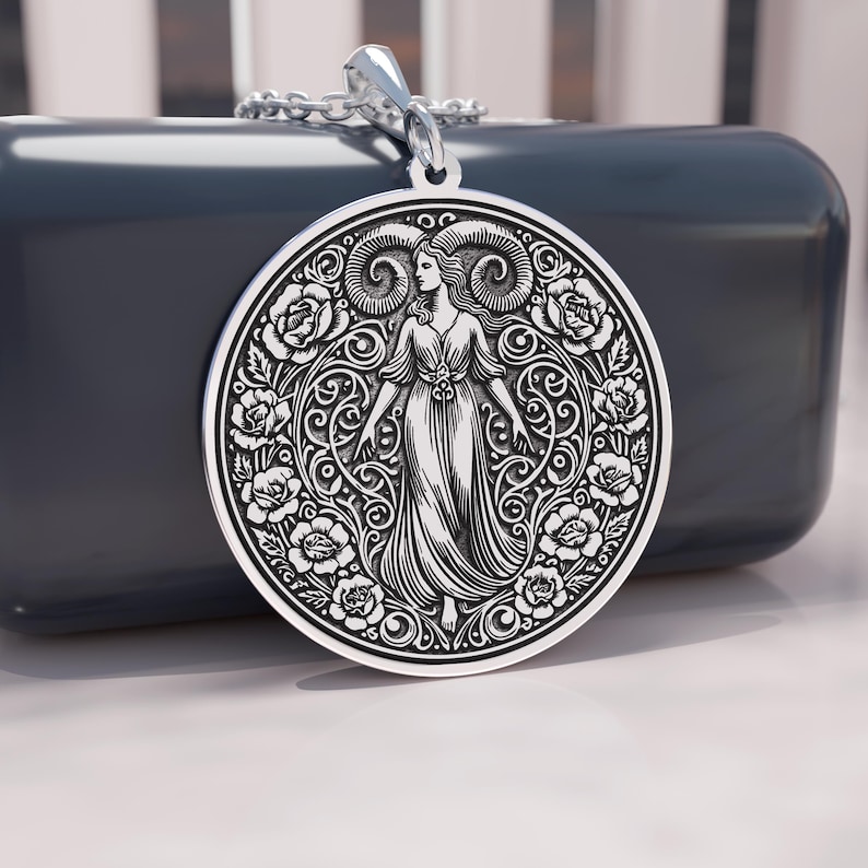 May include: A silver pendant featuring a detailed engraving of a woman with ram horns, surrounded by roses and ornate scrollwork. The pendant hangs from a silver chain. The design is circular and has a vintage aesthetic.