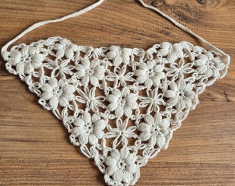 May include: A white, handmade crochet bandana featuring a floral design. The triangular scarf is made of white yarn and has a string tie. The bandana is displayed on a wooden surface.
