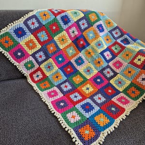 Puede incluir: Una manta de ganchillo hecha a mano compuesta por numerosos cuadrados de la abuela multicolores en tonos rojo, azul, amarillo, verde, naranja y rosa. La manta presenta un borde de color crema con un pequeño fleco decorativo de borlas.