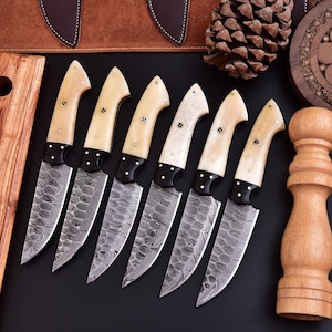 May include: A set of seven Damascus steel knives with cream-colored handles and black accents. The blades have a unique patterned design. The knives are arranged on a black surface, with a wooden cutting board and other kitchen accessories in the background.