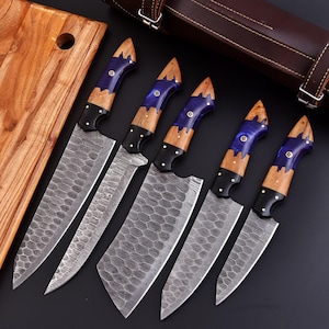 May include: A set of five chef knives with patterned blades and handles featuring wood, blue resin, and black accents. The knives are displayed with a wooden cutting board and a brown leather knife case.