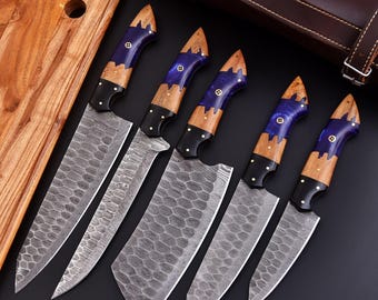 May include: A set of five chef knives with patterned blades and handles featuring wood, blue resin, and black accents. The knives are displayed with a wooden cutting board and a brown leather knife case.