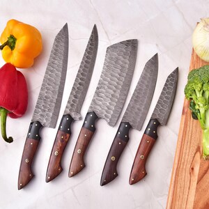 May include: Set of five forged steel kitchen knives with wooden handles. The blades have a textured finish. The handles are dark wood with brass rivets. Red and yellow bell peppers, an onion, and broccoli are also in the image.