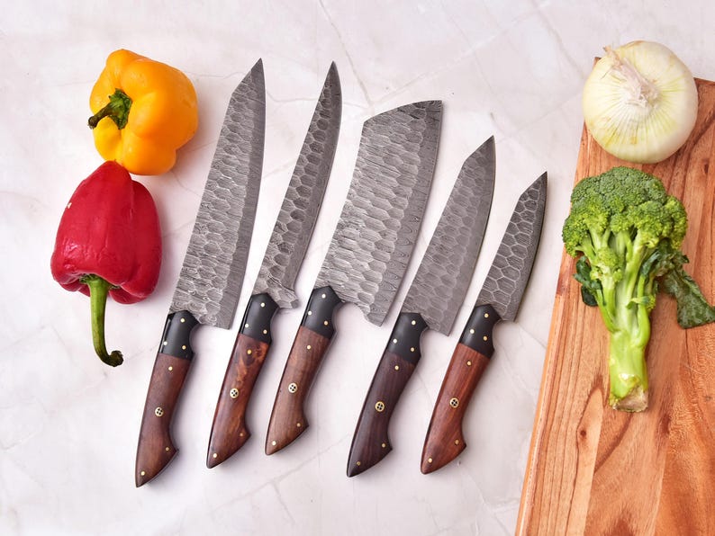 May include: Set of five forged steel kitchen knives with wooden handles. The blades have a textured finish. The handles are dark wood with brass rivets. Red and yellow bell peppers, an onion, and broccoli are also in the image.