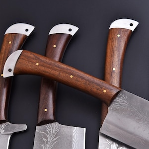 May include: Close-up of several cleavers with dark wooden handles and silver-colored metal accents. The blades have a decorative pattern. The handles are secured with small, gold-colored rivets. The cleavers are arranged against a dark background.