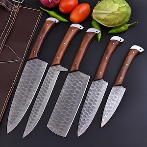 May include: A set of five kitchen knives with wooden handles and textured blades. The knives have a dark brown handle with a light-colored accent at the end. The blades have a unique pattern. A brown leather apron is visible in the background.