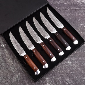 May include: A set of six steak knives presented in a black box. The knives have patterned steel blades, dark brown wooden handles, and silver bolsters. The blades have a serrated edge.