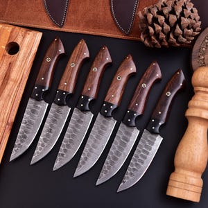 May include: A set of seven kitchen knives with dark wooden handles and patterned blades. The handles are adorned with small, decorative rivets. The blades have a unique, textured design. The knives are arranged on a black surface.