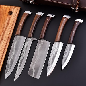 May include: A set of five kitchen knives with wooden handles and patterned blades. The blades have a silver finish with an etched design. The handles are dark brown with brass rivets. A wooden cutting board is visible on the left, and a leather case is in the background.
