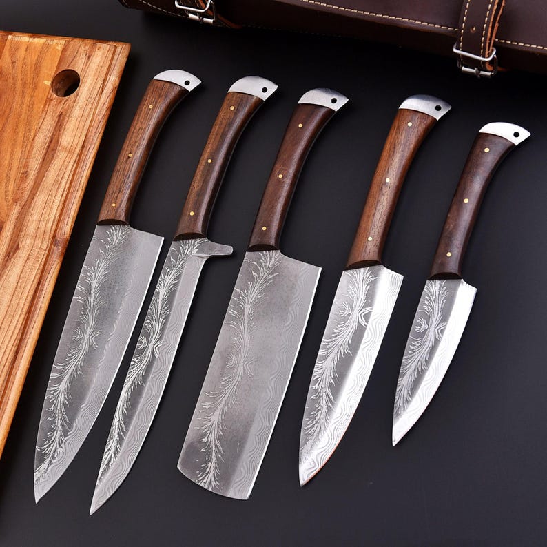May include: A set of five kitchen knives with wooden handles and patterned blades. The blades have a silver finish with an etched design. The handles are dark brown with brass rivets. A wooden cutting board is visible on the left, and a leather case is in the background.