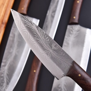 May include: Close-up of a chef's knife with a patterned blade and a dark wooden handle. The blade has a decorative etched design. Other knives are visible in the background, suggesting a set of kitchen knives.