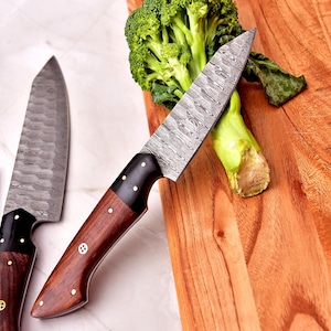 May include: A close-up of several Damascus steel knives with wooden handles. One knife rests on a wooden cutting board next to a head of broccoli. The knives have a patterned blade and dark wood handles with brass rivets.