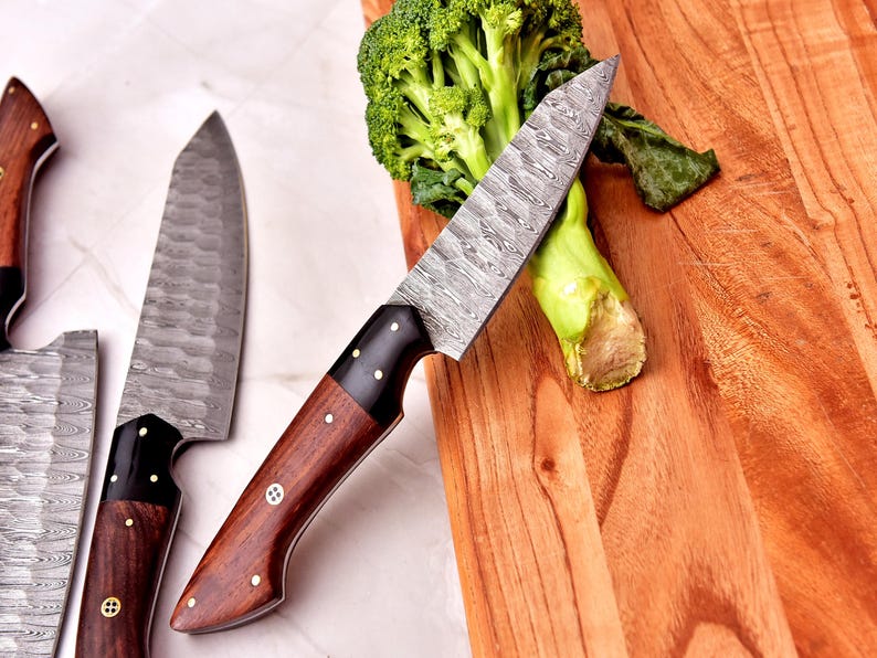 May include: A close-up of several Damascus steel knives with wooden handles. One knife rests on a wooden cutting board next to a head of broccoli. The knives have a patterned blade and dark wood handles with brass rivets.