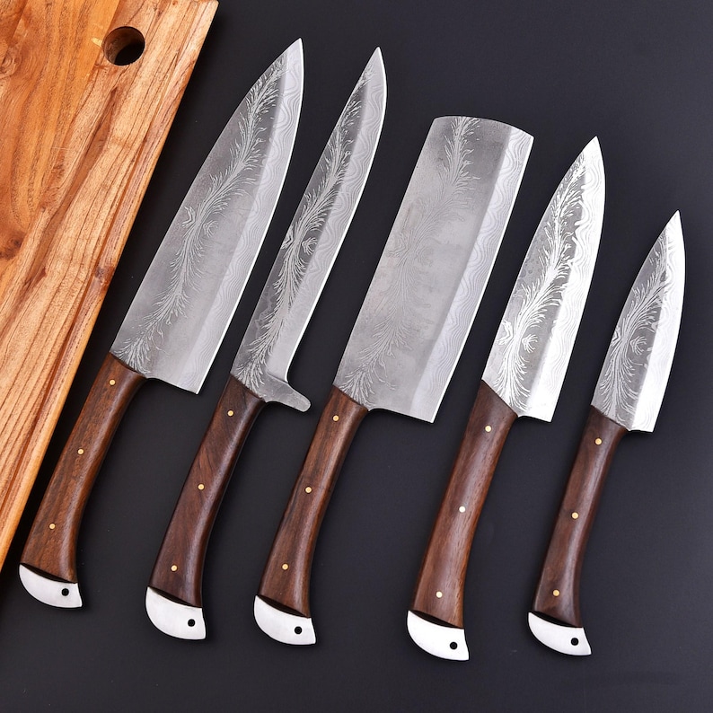 May include: A set of six kitchen knives with dark wooden handles and silver blades. The blades have a decorative etched pattern. The handles are attached to the blades with brass rivets and white metal bolsters. A wooden cutting board is in the background.
