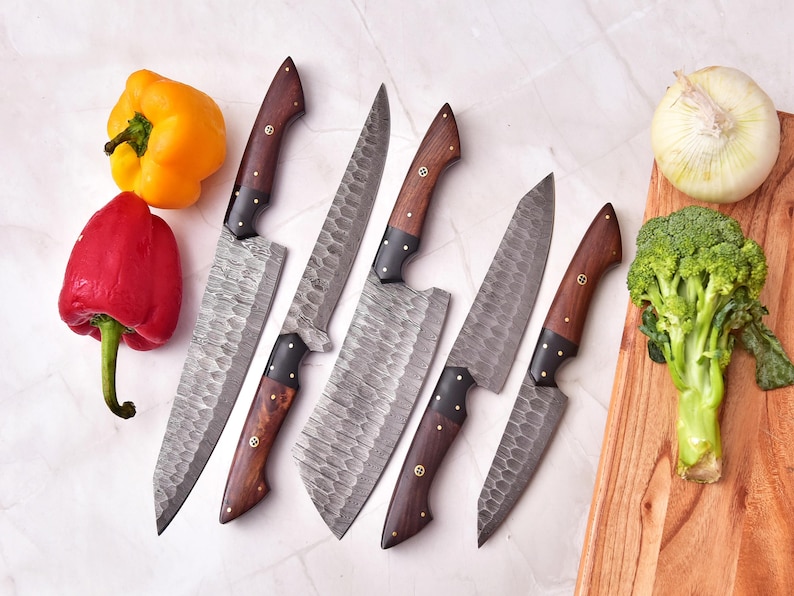 May include: An overhead shot showcases a set of six kitchen knives with dark wooden handles and textured blades. The knives are arranged near fresh produce, including a yellow bell pepper, a red bell pepper, an onion, and a head of broccoli.