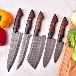 May include: A set of six forged steel kitchen knives with wooden handles and brass rivets. The blades have a textured finish. Yellow and red bell peppers, an onion, and broccoli are arranged with the knives on a white surface and a wooden cutting board.