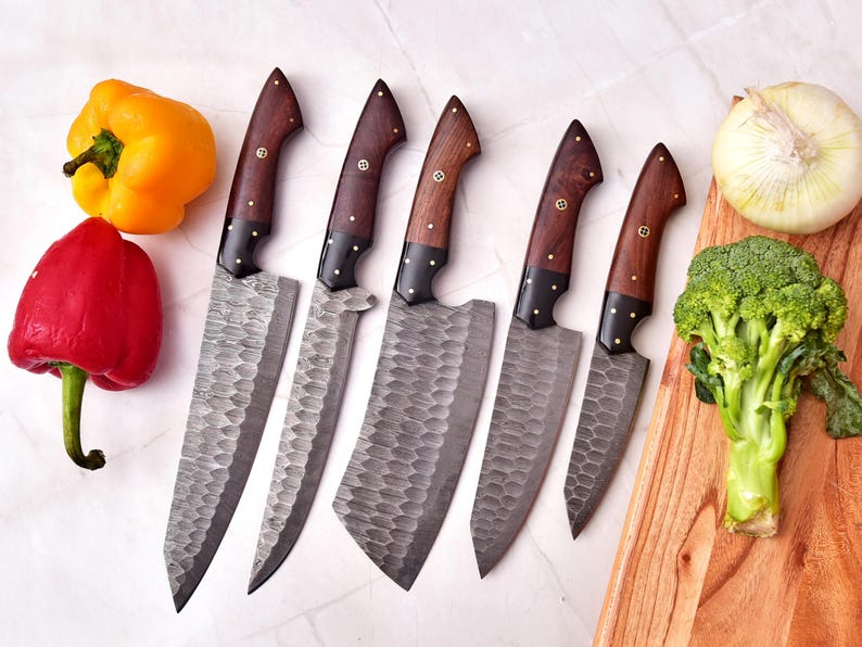 May include: A set of six forged steel kitchen knives with wooden handles and brass rivets. The blades have a textured finish. Yellow and red bell peppers, an onion, and broccoli are arranged with the knives on a white surface and a wooden cutting board.