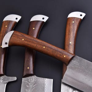 May include: Close-up of several cleavers with dark wooden handles and silver-colored metal accents. The blades have a decorative pattern. The handles are secured with small, gold-colored rivets. The cleavers are arranged against a dark background.