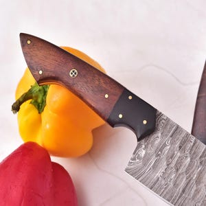 May include: Two Damascus steel chef knives with dark wood handles and brass rivets are displayed. A yellow bell pepper and a red bell pepper are in the background, suggesting a culinary theme.