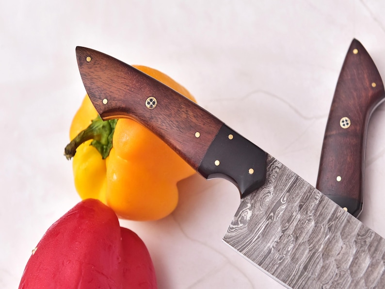 May include: Two Damascus steel chef knives with dark wood handles and brass rivets are displayed. A yellow bell pepper and a red bell pepper are in the background, suggesting a culinary theme.