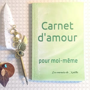 Könnte beinhalten: Ein hellgrünes Notizbuch mit den Worten "Carnet d'amour pour moi-même" in Grün. Ein dekorativer Stift mit weißer Feder, Zahnrädern und goldfarbener Feder neben dem Notizbuch. Das Notizbuch ist von "Les carnets de Naëlle."