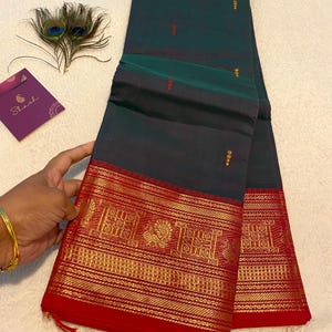 May include: Dark teal saree with a red and gold border featuring intricate patterns. The saree is folded, showcasing the rich colours and detailed design. A peacock feather and a purple card are in the background.