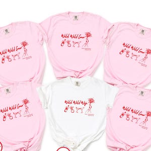 May include: Five short-sleeved t-shirts arranged in a group. Four shirts are light pink and one is white. Each shirt features a red graphic print with the text Wild Wild Crew or Wild Wild Wife, accompanied by illustrations of a bottle, ring, and glass.