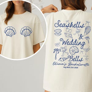 May include: Cream-coloured t-shirt with a blue seashell design on the front and a back graphic that reads "Seashells & Wedding Bells, Olivia's Bachelorette, Key West, Est: 2026". The shirt has a relaxed, oversized fit.